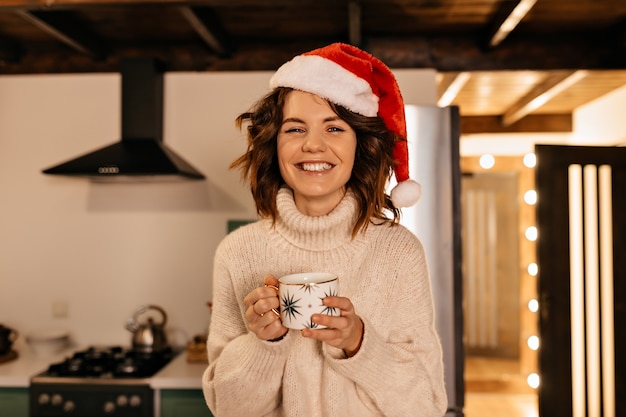 Adorable jolie femme aux cheveux bouclés portant des vêtements tricotés et bonnet de Noel assis sur la cuisine et en attente de fête de Noël