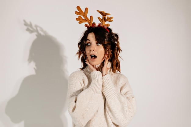 Adorable jolie fille avec des boucles portant un accessoire de tête et un pull beige regardant loin avec des émotions surprises et une bouche ouverte sur fond isolé