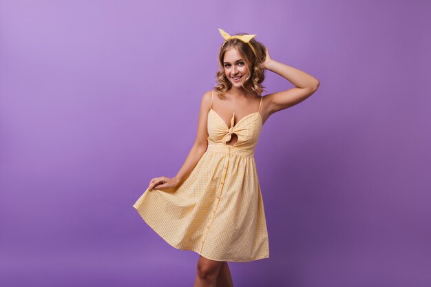 Adorable modèle féminin en robe à carreaux jaune dansant avec le sourire. portrait de jeune fille blonde spectaculaire bénéficiant d'une bonne journée.