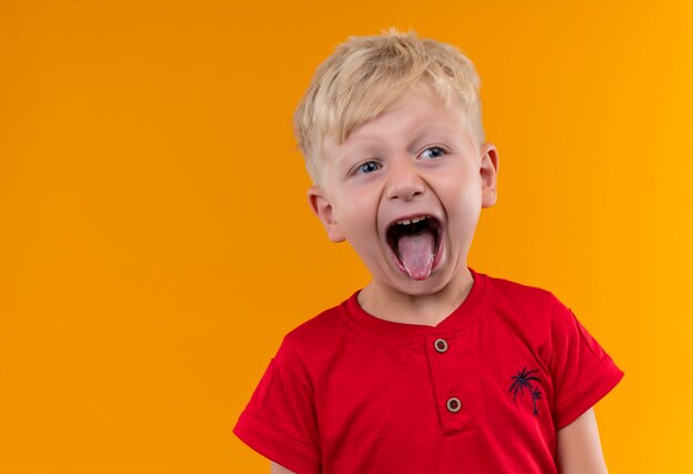 Un adorable petit garçon aux cheveux blonds et aux yeux bleus portant un t-shirt rouge montrant sa langue tout en regardant de côté