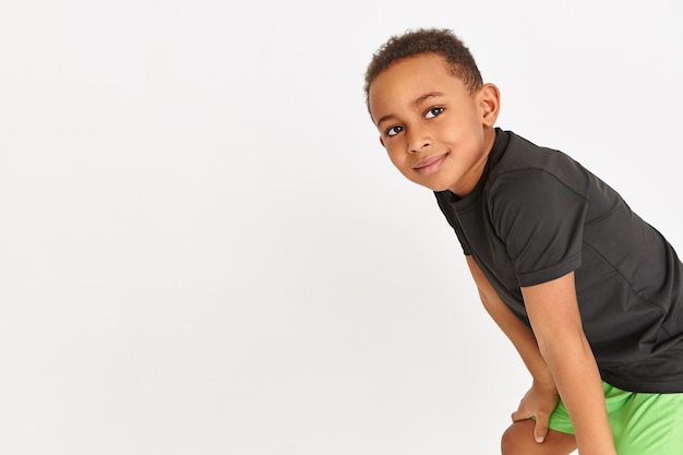 Adorable petit garçon en t-shirt noir et short vert au repos pendant l'entraînement cardio en gardant les mains au-dessus de ses genoux