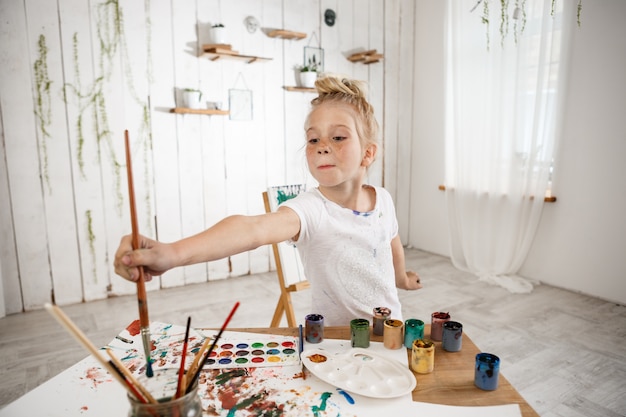 Photo gratuite adorable petite fille caucasienne créative et talentueuse tenant un gros pinceau dans sa main et l'enfonçant dans l'eau, debout derrière le bureau dans la salle d'art.