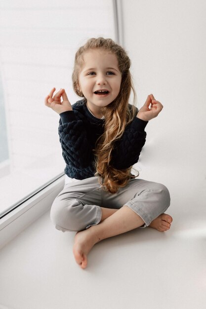 Adorable petite fille charmante aux longs cheveux ondulés est assise sur la fenêtre et lève les mains avec un sourire charmant et heureux