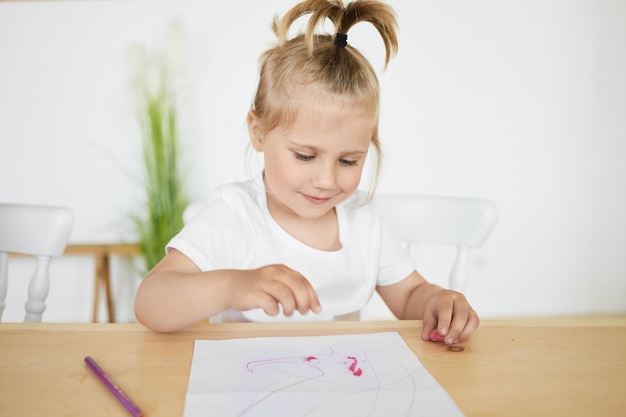 Adorable petite fille charmante avec queue de cheval assis au bureau à la maternelle devant une feuille blanche, colorant ou faisant des figures à l'aide de pâte à modeler ou d'argile, ayant une expression faciale joyeuse et heureuse