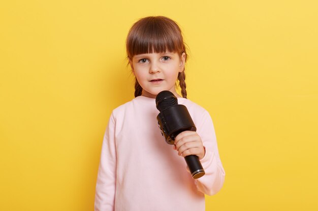 Adorable petite fille avec microphone sur fond jaune, regarde la caméra tout en parlant au micro, pointant l'index de côté. Copiez le rythme pour la publicité ou le texte promotionnel.