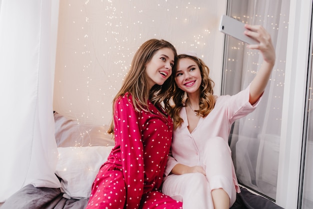 Adorables filles assises sur le lit et posant pour selfie. Photo intérieure de deux jeunes femmes détendues appréciant une bonne matinée ensemble.