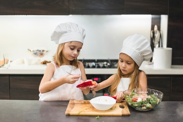 Adorables soeurs mignonnes en toque et tabliers préparant un repas en cuisine