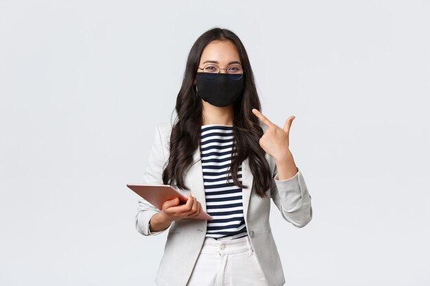 Affaires, finances et emploi, covid-19 empêchant le virus et concept de distanciation sociale. Femme d'affaires asiatique souriante avec tablette numérique pointant sur un masque de protection sur le visage