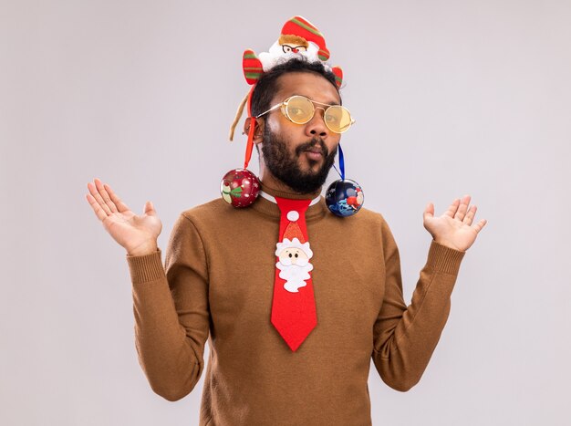 African American man in brown pull et santa rim sur la tête avec une cravate rouge drôle avec des boules de Noël sur ses oreilles à la levée confus des bras debout sur fond blanc