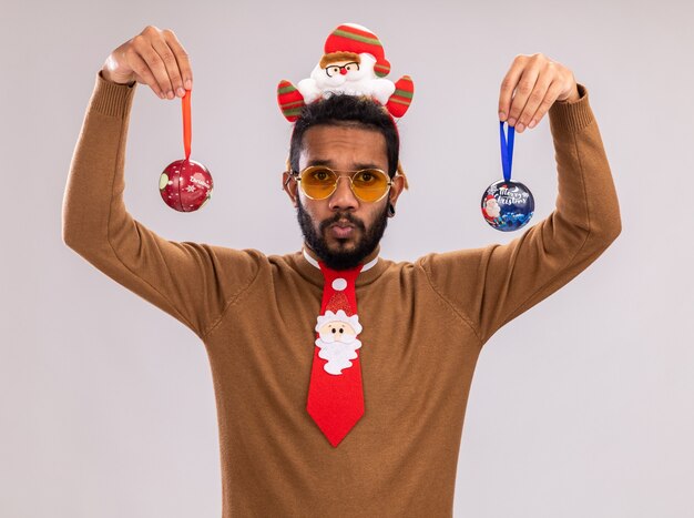 African American man in brown pull et santa rim sur la tête avec une cravate rouge drôle tenant des boules de Noël avec une expression de confusion debout sur un mur blanc