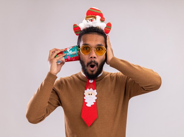 African American man in brown pull et santa rim sur la tête avec une cravate rouge drôle tenant une tasse de papier coloré sur son oreille à la stupéfaction debout sur fond blanc