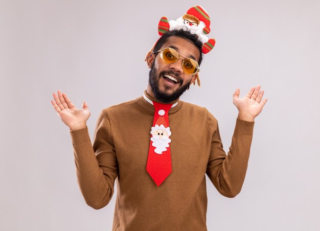 African American man in brown pull et santa rim sur la tête avec drôle cravate rouge regardant la caméra souriant joyeusement debout sur fond blanc