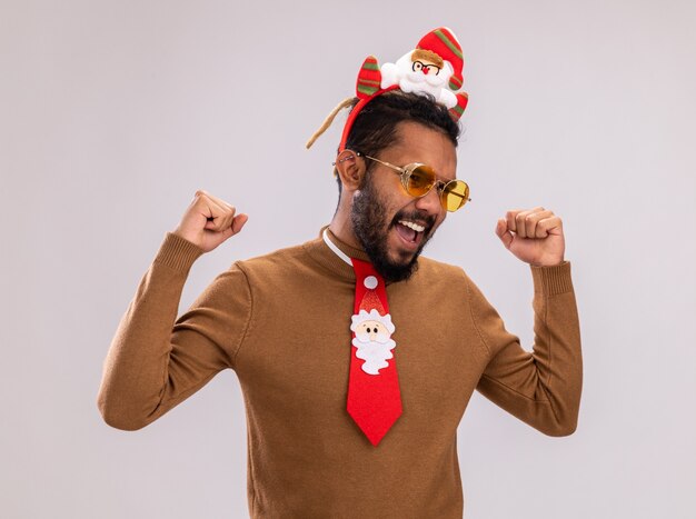 African American man in brown pull et santa rim sur la tête avec drôle cravate rouge serrant les poings heureux et excité debout sur fond blanc