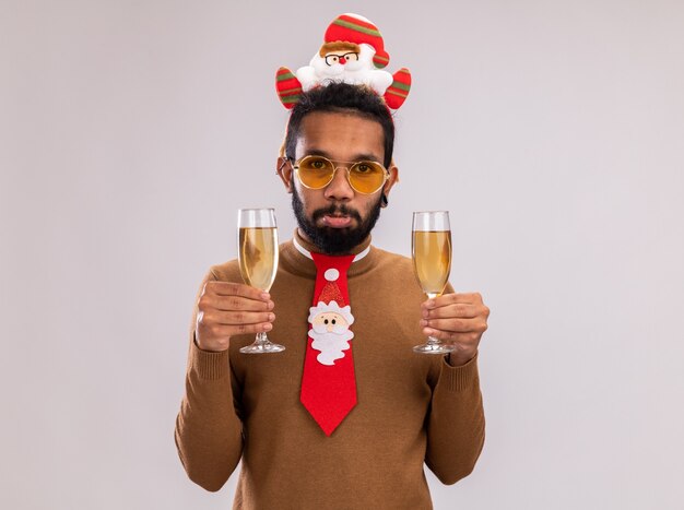 African American man in brown pull et santa rim sur la tête avec drôle cravate rouge tenant deux verres de champagne regardant la caméra avec une expression triste debout sur fond blanc