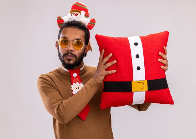 African American man in brown pull et santa rim sur la tête avec drôle cravate rouge tenant un oreiller de Noël regardant la caméra souriant confiant debout sur fond blanc