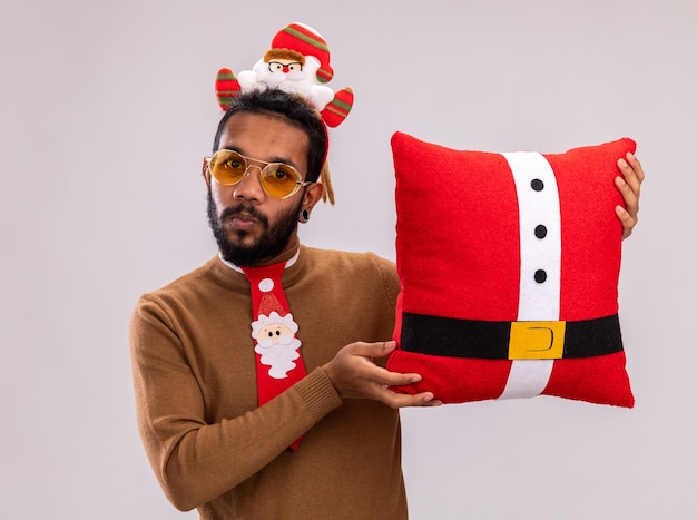 African American man in brown pull et santa rim sur la tête avec drôle cravate rouge tenant un oreiller de Noël surpris debout sur un mur blanc