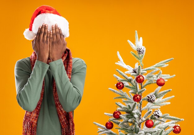 African American man in santa hat et écharpe autour du cou couvrant le visage avec les mains debout à côté d'un arbre de Noël sur mur orange
