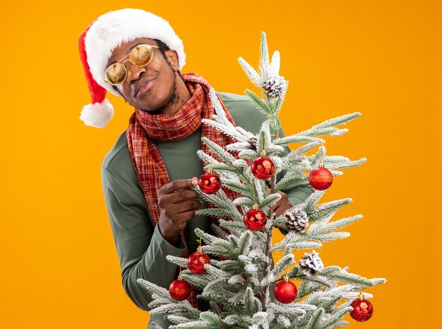 African American man in santa hat et écharpe autour du cou avec sourire sur le visage debout à côté d'un arbre de Noël sur mur orange