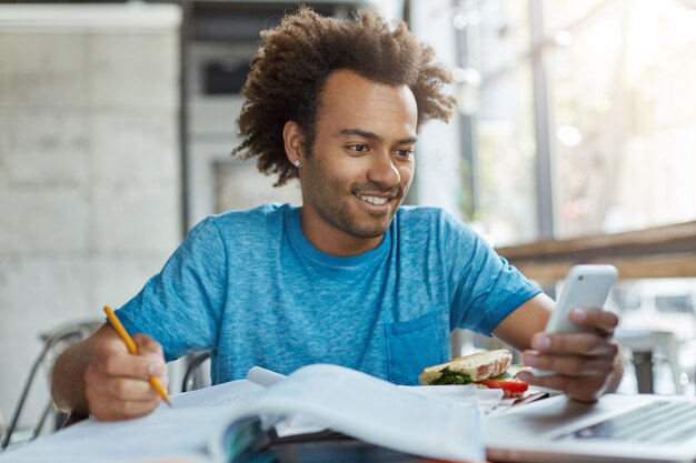 Afro-américain étudiant heureux étant à la cafétéria entouré de livres et de copies de livres se préparant pour les cours en tapant un message texte sur un gadget électronique souriant agréablement en lisant des sms