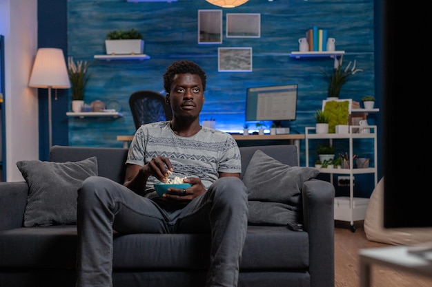 Un afro-américain se reposant sur un canapé dans le salon tenant un bol de pop-corn tout en regardant un film de divertissement à la télévision. jeune homme appréciant de passer du temps libre seul dans le salon
