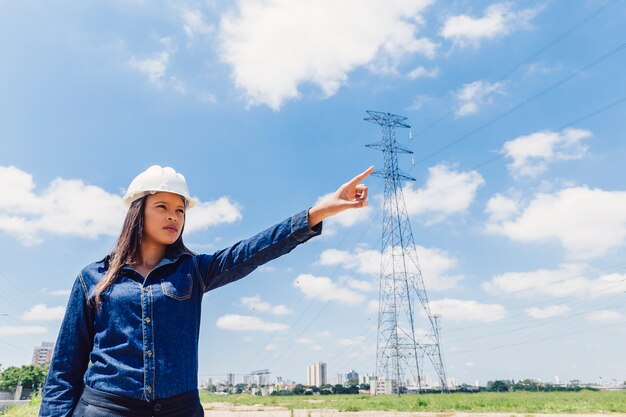 Afro-américaine dans un casque de sécurité pointant près de la ligne à haute tension