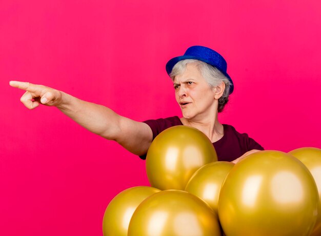 Agacé femme âgée portant chapeau de fête se dresse avec des ballons d'hélium à la recherche et pointant sur le côté rose