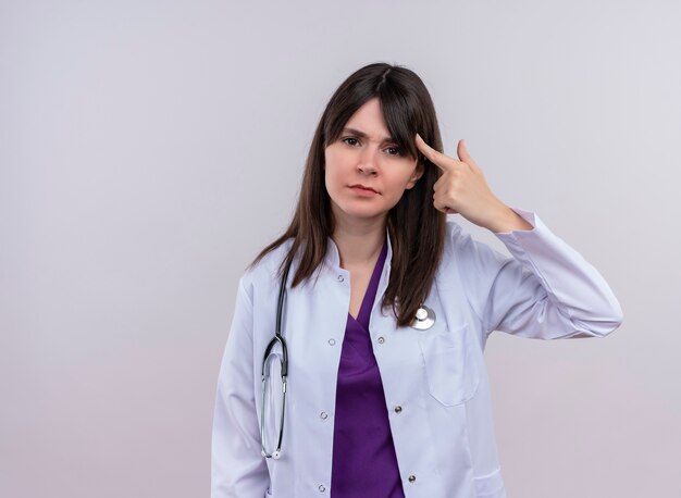 Agacé jeune femme médecin en robe médicale avec stéthoscope met le doigt sur sa tempe sur fond blanc isolé avec copie espace