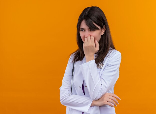 Agacé jeune femme médecin en robe médicale avec stéthoscope met la main sur son menton sur fond orange isolé avec copie espace