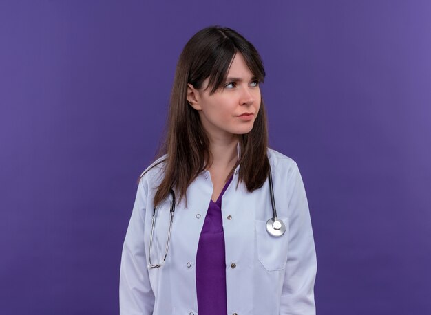 Agacé, jeune femme médecin en robe médicale avec stéthoscope regarde sur le côté sur fond violet isolé avec copie espace