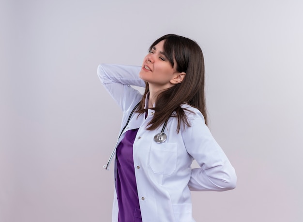 Agacé jeune femme médecin en robe médicale avec stéthoscope se tient sur le côté et tient à l'arrière avec les deux mains sur fond blanc isolé avec copie espace