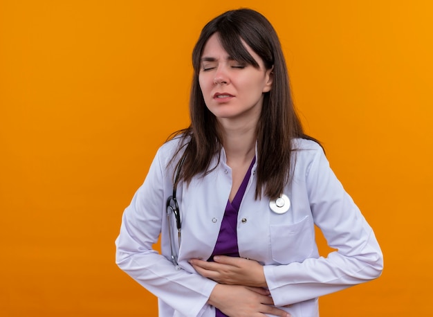 Agacé jeune femme médecin en robe médicale avec stéthoscope tient le ventre à deux mains sur fond orange isolé avec copie espace