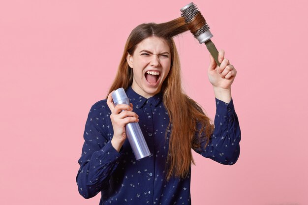 Agacé jeune femme se coiffe les cheveux, insatisfaits du shampooing, tient la brosse à cheveux et la laque, regarde avec une expression irritée à la caméra