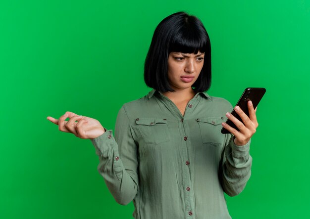 Agacé, jeune fille brune caucasienne regarde le téléphone et tient la main ouverte isolé sur fond vert avec espace copie