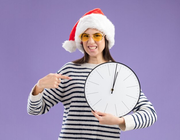 Agacé jeune fille caucasienne dans des lunettes de soleil avec bonnet de Noel tenant et pointant sur l'horloge