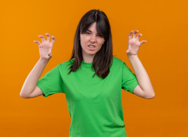 Agacé jeune fille de race blanche en chemise verte a levé les mains et regarde la caméra sur fond orange isolé