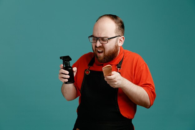 Agacé jeune homme coiffeur portant des lunettes chemise rouge et tablier de coiffeur tenant un fixatif et un peigne regardant le côté isolé sur fond bleu