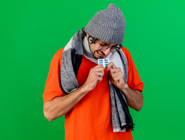 Photo gratuite agacé jeune homme malade portant des lunettes chapeau d'hiver et écharpe tenant pack de capsules médicales avec pack de capsules sous chapeau isolé sur mur vert
