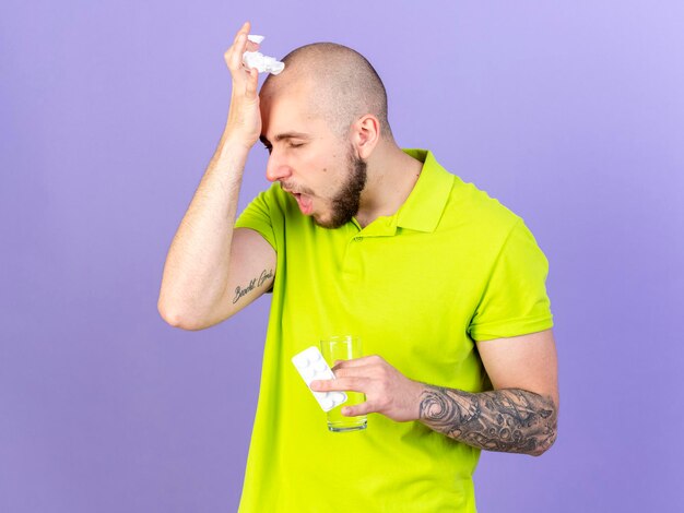 Agacé jeune homme malade de race blanche met la main sur le front détient un verre d'eau et pack de pilules médicales sur violet