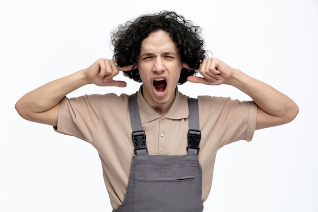 Photo gratuite agacé jeune homme travailleur de la construction portant l'uniforme regardant la caméra mettant les doigts dans les oreilles criant isolé sur fond blanc