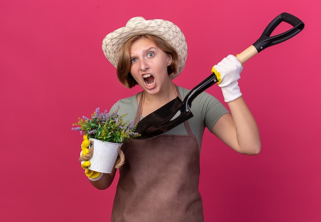 Agacé jeune jardinier femme slave portant chapeau de jardinage et gants tenant des fleurs en pot de fleurs et pelle sur rose