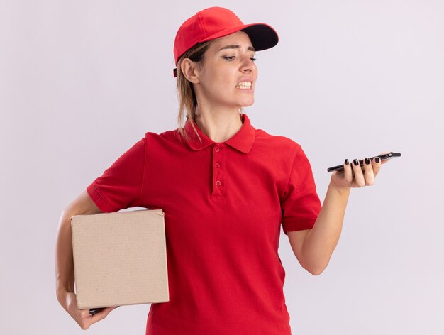 Agacé jeune jolie livreuse en uniforme détient cardbox et regarde le téléphone sur blanc