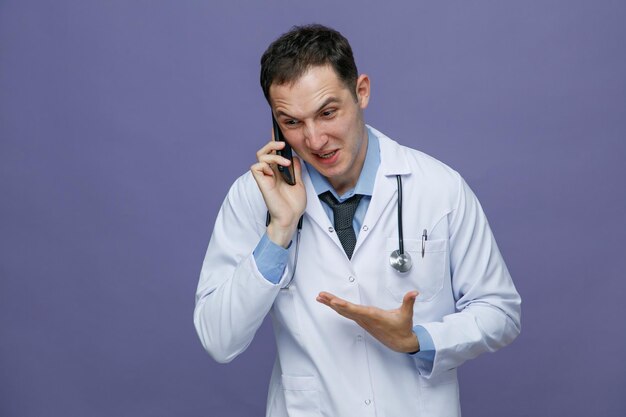 Agacé jeune médecin de sexe masculin portant une robe médicale et un stéthoscope autour du cou montrant une main vide regardant à côté tout en parlant au téléphone isolé sur fond violet