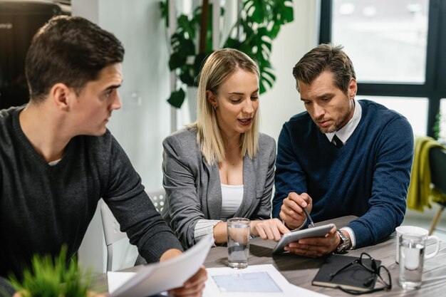 Agent immobilier arrière et jeune couple examinant des plans de logement sur un pavé tactile tout en ayant une réunion à la maison