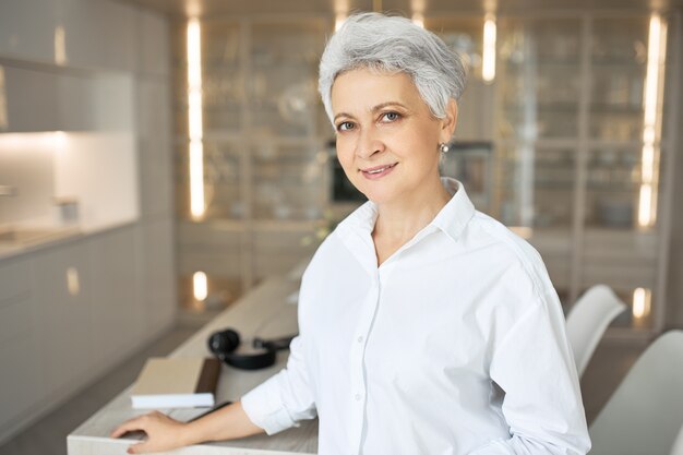 Agent immobilier féminin aux cheveux gris à la mode en chemise blanche posant à l'intérieur. Gestionnaire de femme élégante heureuse debout au bureau pendant la journée de travail