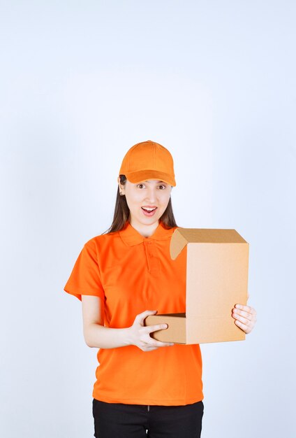 Agent de service féminin en code vestimentaire de couleur orange tenant une boîte en carton ouverte, regarde à l'intérieur et est surpris