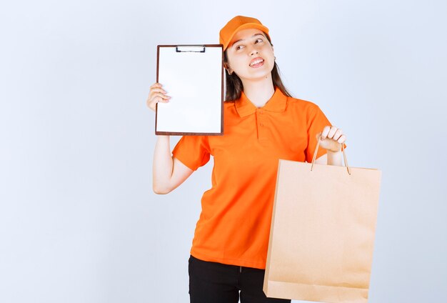 Agent de service féminin en code vestimentaire de couleur orange tenant un sac en carton et présentant la liste de signatures au client tout en ayant l'air réfléchi.