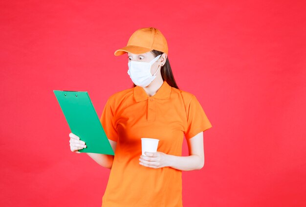 Agent de service féminin en code vestimentaire et masque de couleur orange présentant un nouveau gobelet jetable de marque et vérifiant les détails avec émotions.