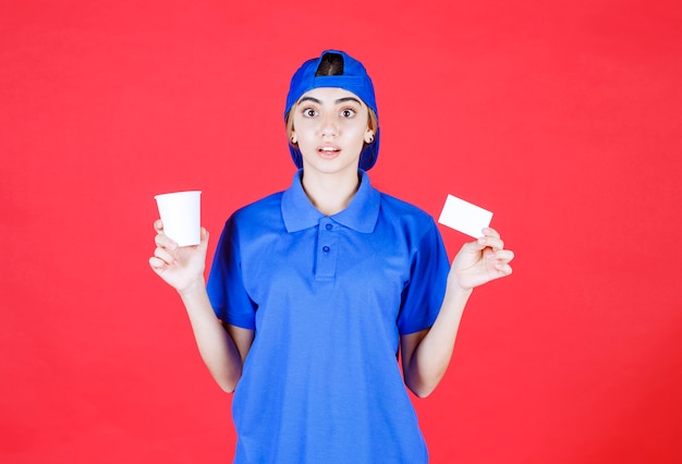 Agent de service féminin en uniforme bleu tenant une tasse de boisson et présentant sa carte de visite tout en l'air surpris.