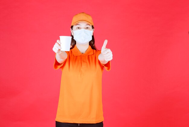 Agent de service féminin en uniforme de couleur orange et masque tenant un gobelet jetable et montrant un signe de main positif.