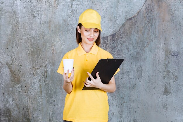 Agent de service féminin en uniforme jaune tenant un gobelet en plastique et un dossier d'adresses noir.